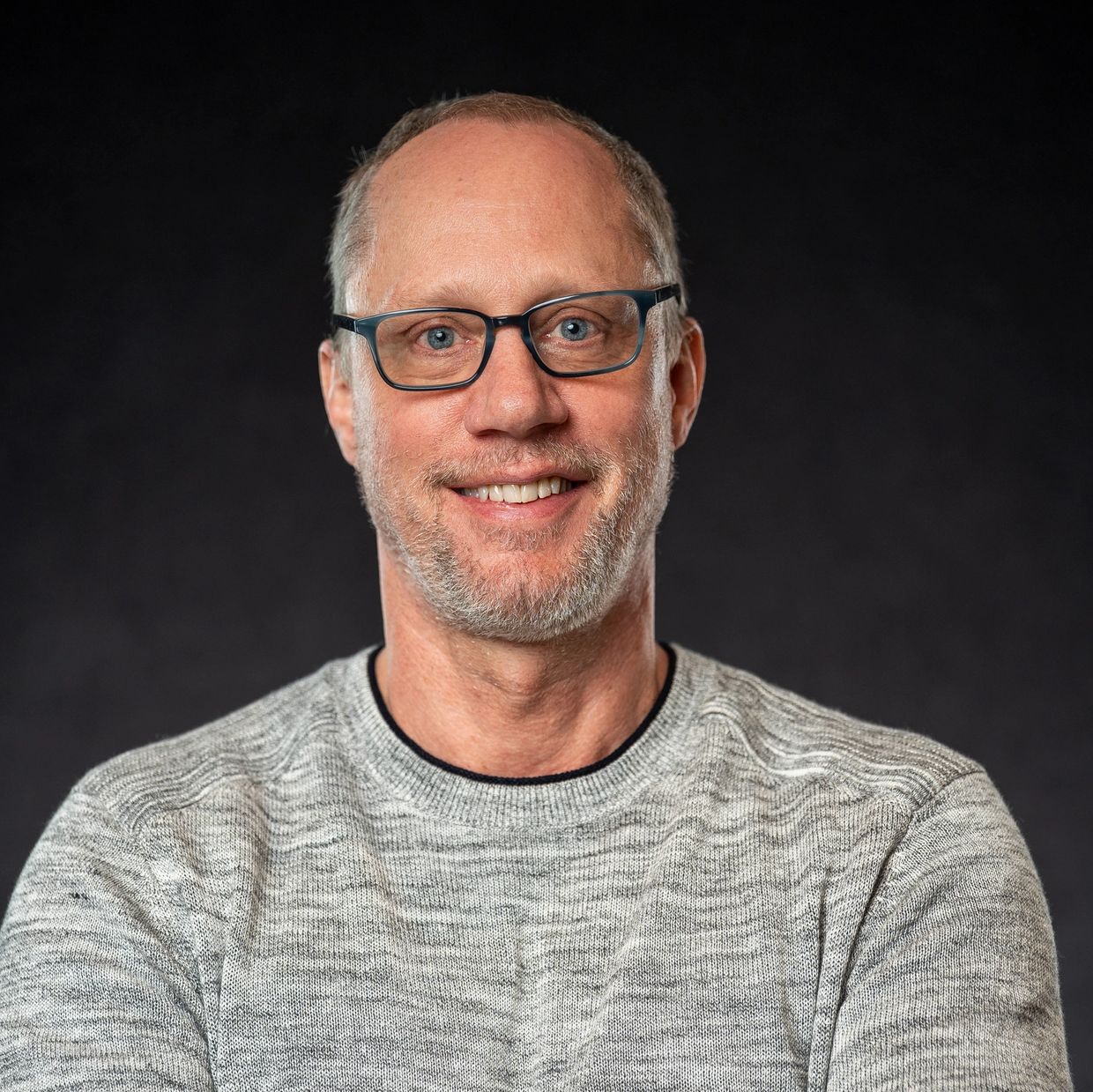 Smiling man with glasses wearing a grey sweater against a dark background.