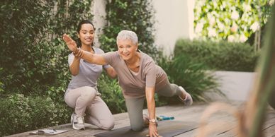 Personal trainer assists elderly woman with balance exercise on yoga mat outdoors.