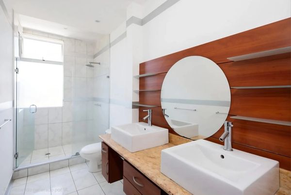 Modern bathroom with double sinks, round mirror, and glass shower enclosure.