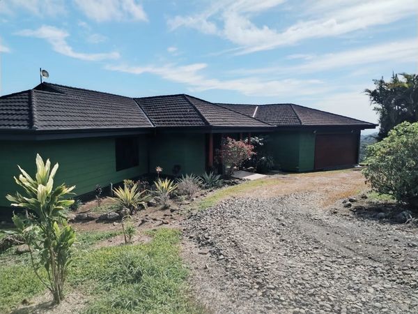 Single-story green house with a gravel driveway and tropical plants under a blue sky.