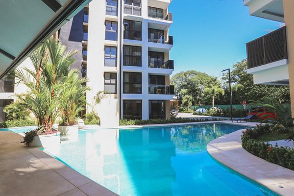 Modern apartment complex with a clear blue swimming pool and lush greenery.