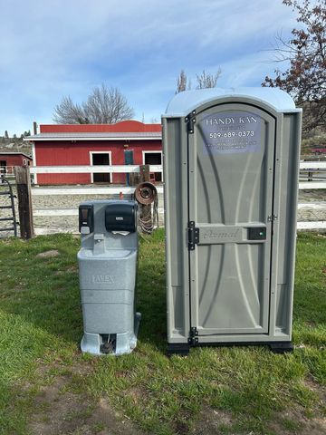 Portable toilet and hand sanitizer station on a grassy area near a red barn.