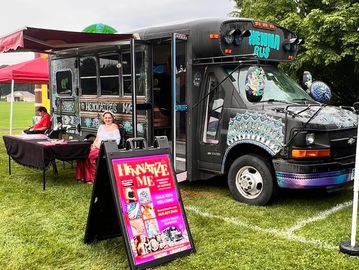 The Hennatize Me Henna Bus set up at an outdoor event with awning, table display, and signage.
