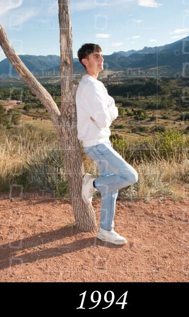 Young man leaning against a tree outdoors with mountains in the background.