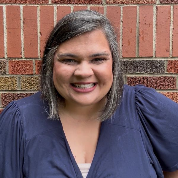 Smiling woman with gray-streaked hair in a navy blue top against a brick wall.