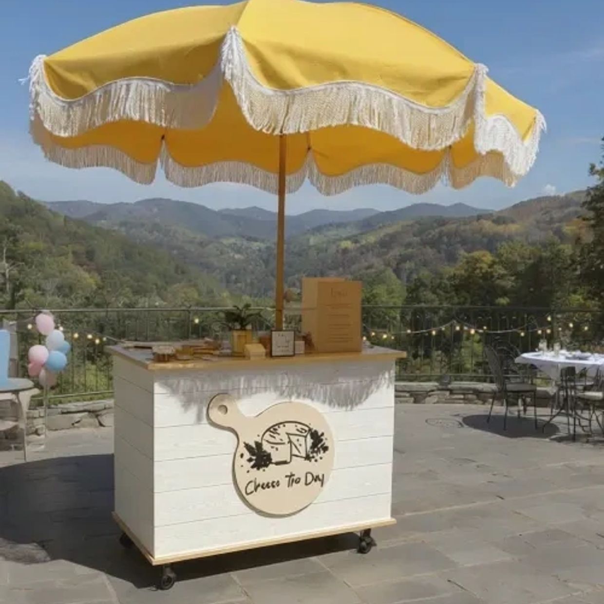 Outdoor cheese stand with yellow fringed umbrella and mountain view.