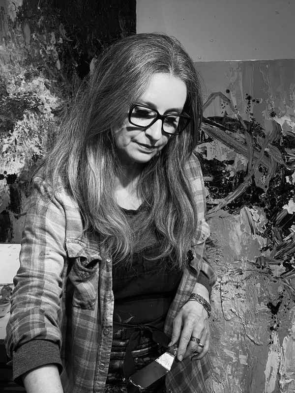 Woman artist with glasses holding a palette knife in front of abstract paintings.