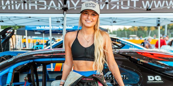 Woman racer smiling beside her race car, wearing a cap and racing suit.