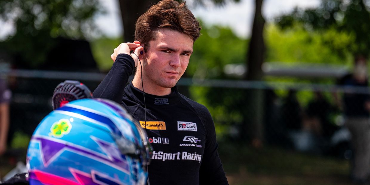 Race car driver adjusting earphones with helmet in foreground.