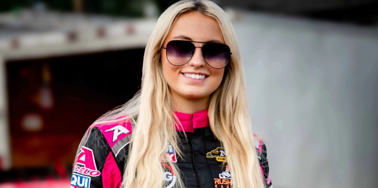 Smiling female race car driver with long blonde hair and sunglasses.