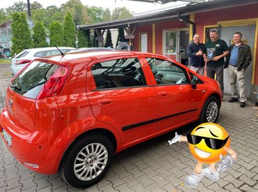 Trzech uśmiechniętych mężczyzn stoi przed czerwonym samochodem Fiat Punto  + emotion w okularach