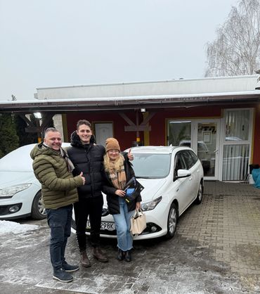 Dwóch męzczyzn i obejmowana kobieta stoją przy białej Toyotcie Auris kombi, Jeden pokazuje kciuk OK.