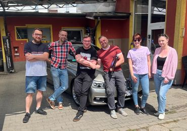 Grupka młodych uśmiechniętych stoi przed samochodem i dwóch podaje sobie ręce