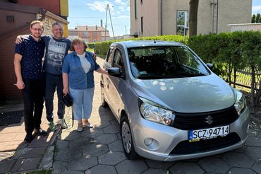 trzy osoby stoją obok srebrnego Suzuki Celerio. Mężczyzna po środku obejmuje kobietę i mężczyznę.