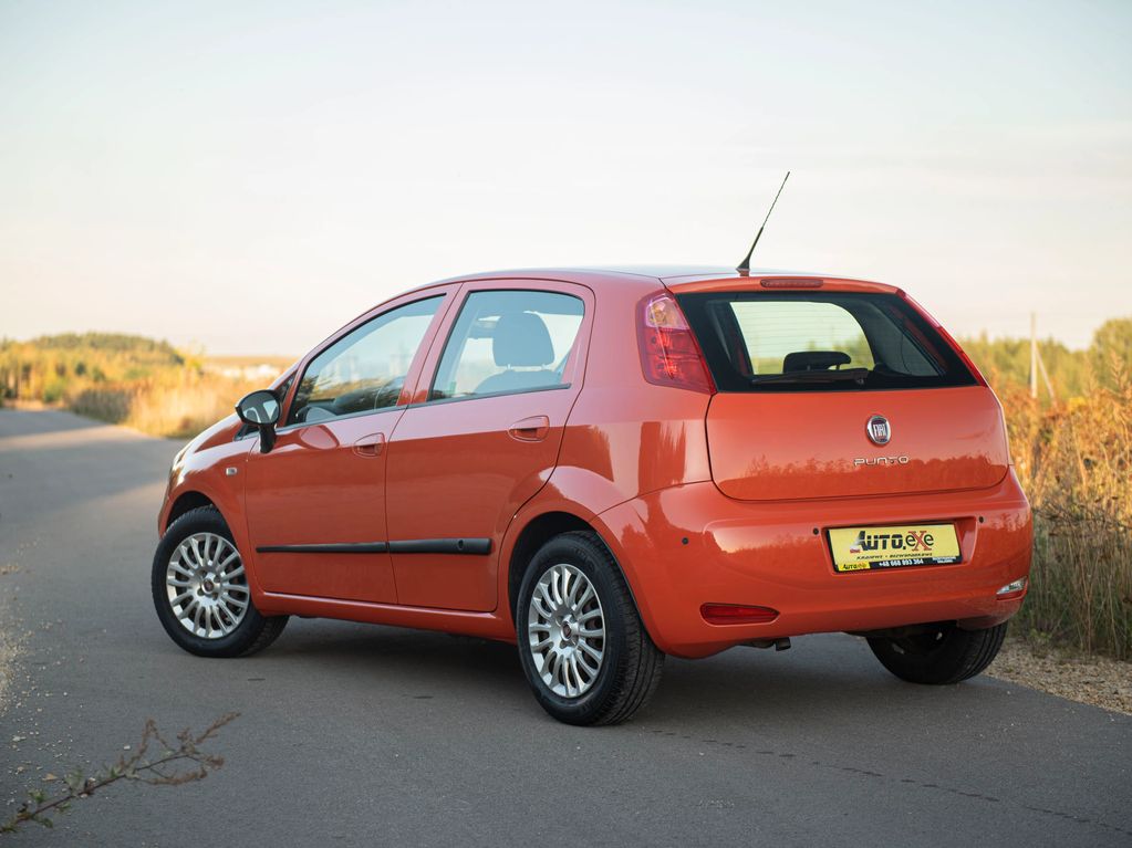 Fiat Punto w kolorze marchewkowym stoi skosem na wąskiej drodze asfaltowej w plenerze lewym tyłem