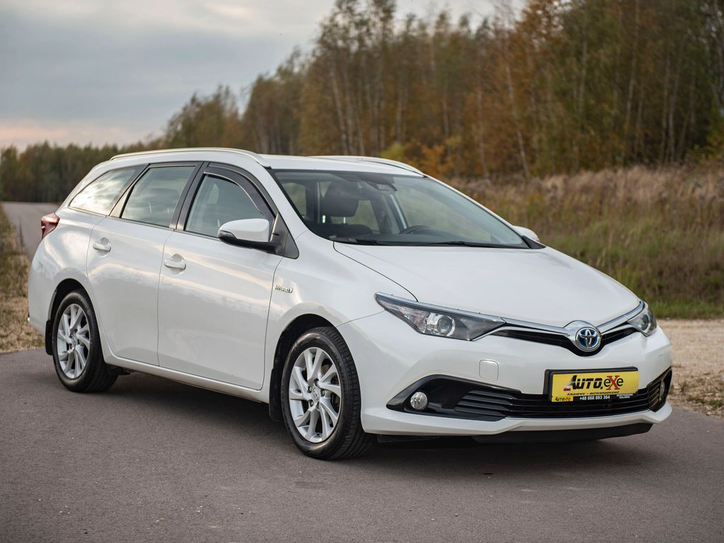 Toyota Auris kolor biały stoi przednim prawym bokiem na drodze asfaltowej obok trawy