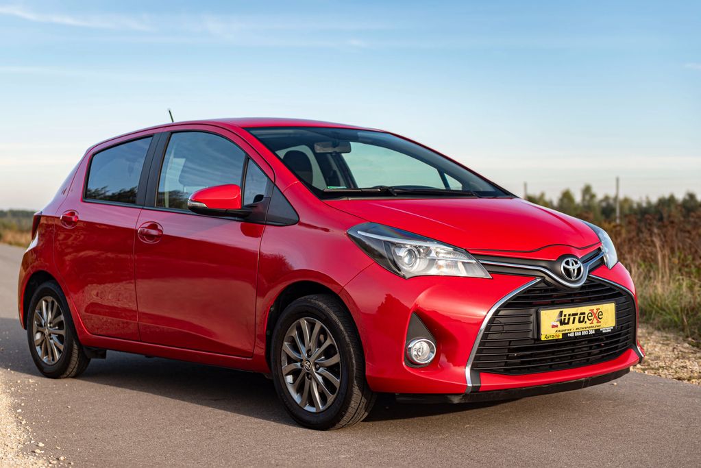 toyota Yaris kolor czerwony stoi przednim prawym bokiem na drodze asfaltowej obok trawy