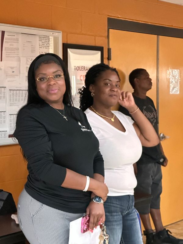 Three people standing indoors against an orange wall, two women smiling at the camera.