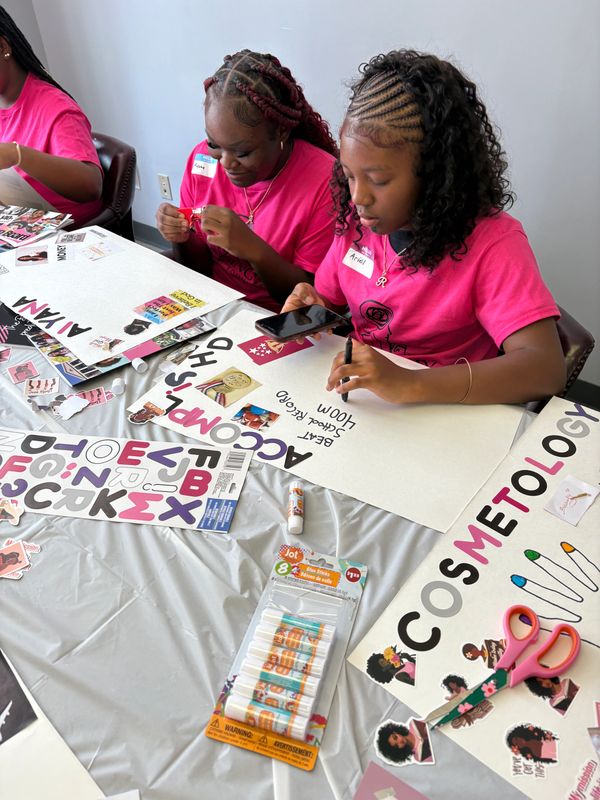 Two girls in pink shirts making creative posters with stickers and glue sticks.