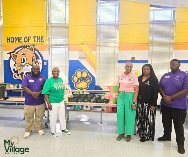 Group of five people standing in a school cafeteria with donation boxes and colorful wall art.