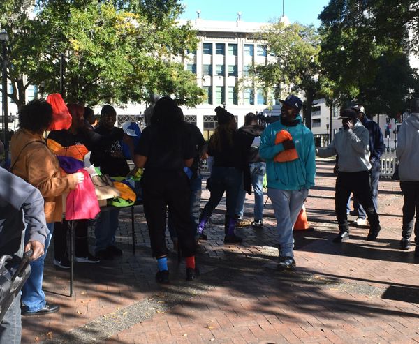 People gather outdoors exchanging colorful bags on a sunny day.