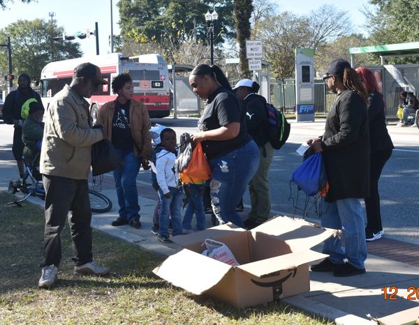 Community members gather outdoors to distribute backpacks and supplies near a bus stop.