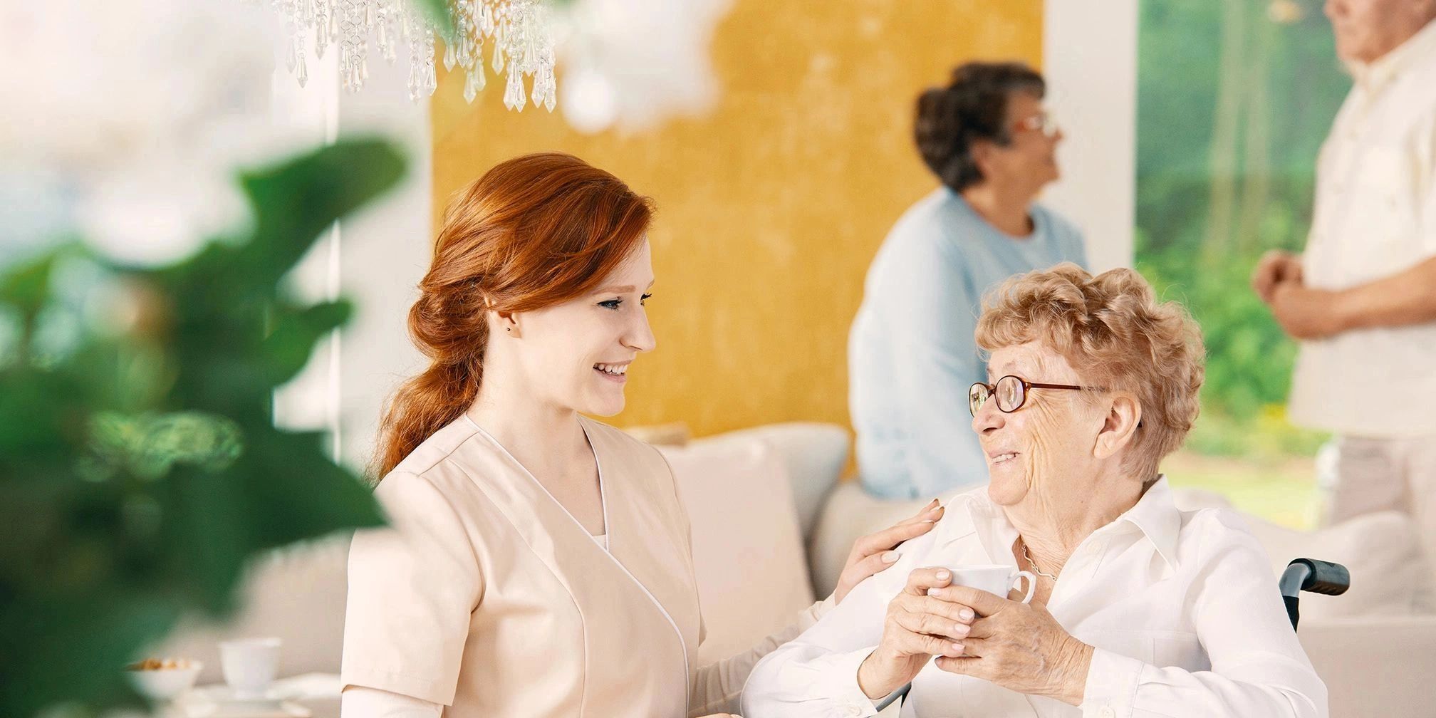 Caregiver smiling warmly at an elderly woman holding a cup.