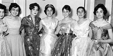 Seven women in elegant vintage dresses posing together indoors.
