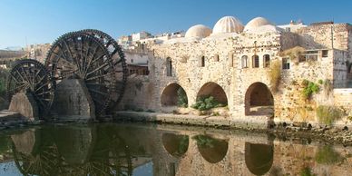 Ancient water wheels beside historic stone buildings reflected in calm water.