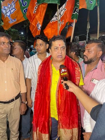 Man in yellow shirt with red scarf being interviewed with BJP flags in background.