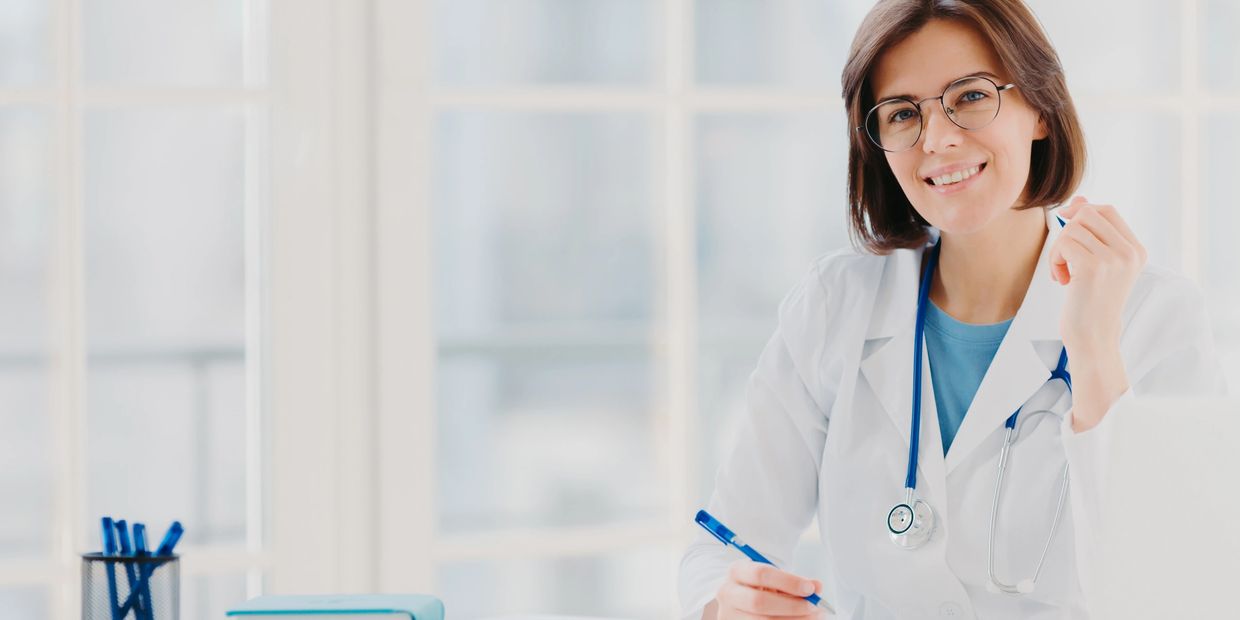 Smiling female doctor with glasses and stethoscope at desk with books and laptop.