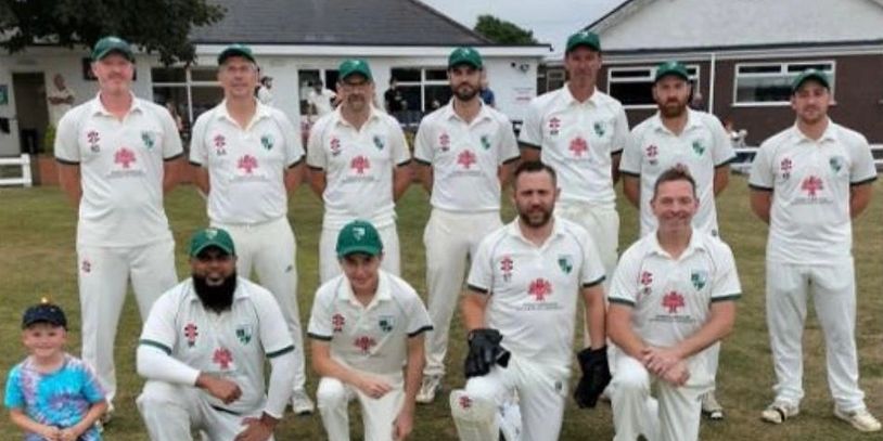 Cricket team posing outdoors in white uniforms with a young child.