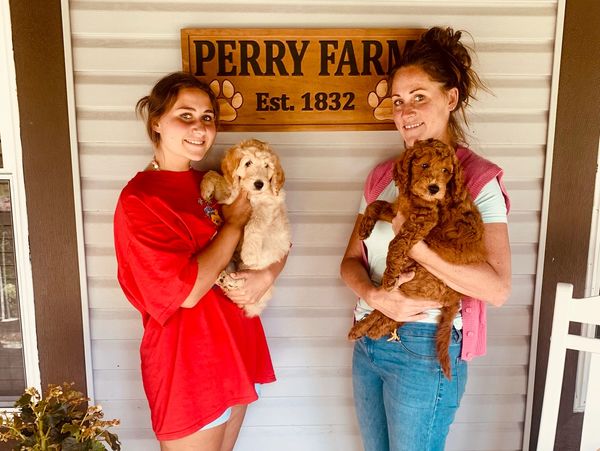 Two women smiling and holding puppies on a porch out front of a sign that reads Perry Farm Est 1832.