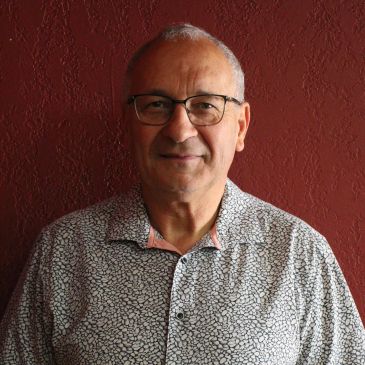 Man wearing glasses and a patterned shirt standing against a red wall.