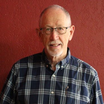 Elderly man wearing glasses and a plaid shirt against a red wall.