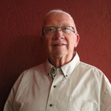 Elderly man with glasses smiling against a red wall.