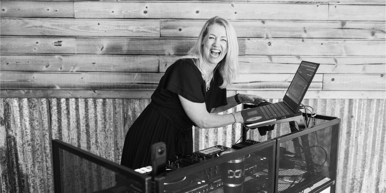 A joyful woman DJ laughs while working on a laptop and DJ equipment.