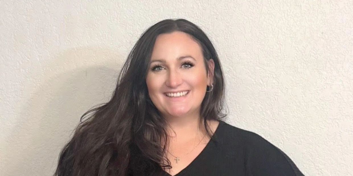 Smiling woman with long dark hair wearing a black top against a textured white wall.