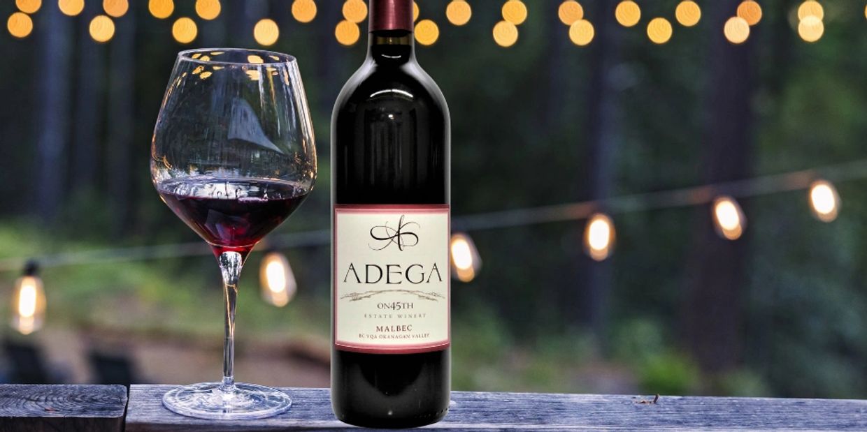 Bottle of Adega Malbec wine and a filled glass on a wooden surface with blurred lights in the background.