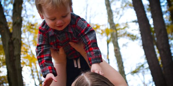 A woman lifts a smiling toddler outdoors among tall trees with autumn leaves.