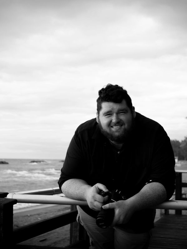 Smiling man holding a camera on a wooden railing by the beach.