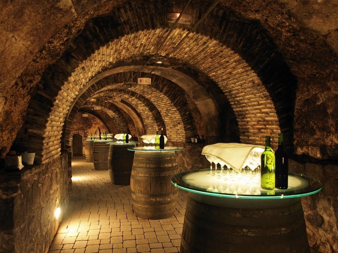 Wine cellar with barrels and glasses under arched brick ceilings.
