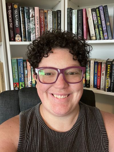Smiling person with curly hair and purple glasses in front of a bookshelf.
