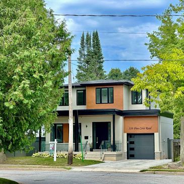 Modern two-story house with large windows and a garage, surrounded by lush green trees.