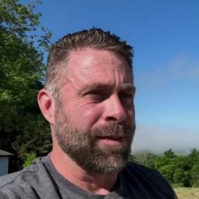 Bearded man outdoors with trees and blue sky.