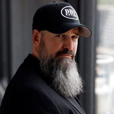 Man with a long gray beard wearing a black cap and jacket.