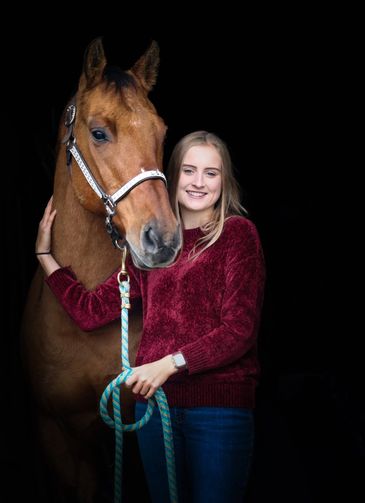 Young woman in a red sweater poses with a brown horse against a dark background.