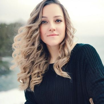 Michelle wearing black sweater with curly, long, blonde hair with a blurred background.  