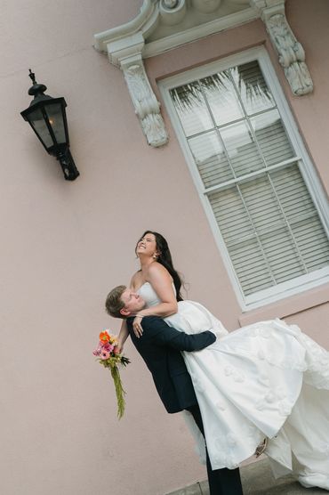 Groom lifting bride joyfully outside a pastel pink building.