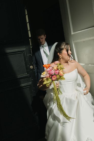 Bride in white gown holding bouquet, groom behind in suit by dark door.